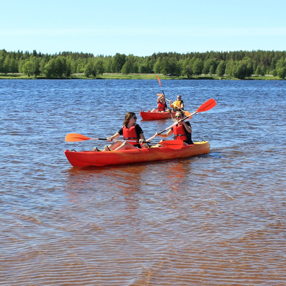 2 människor som paddlar i en kajak på vattnet nära strandkanten. I bakgrunden syns en till kajak med två människor, och längre bort en grön skog med blå himmel.