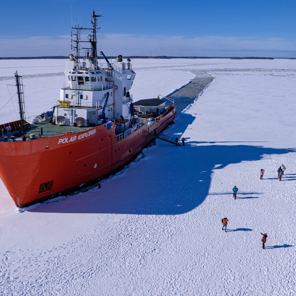 Stort rött fartyg som brutit en ränna i isen. Människor går på isen bredvid.