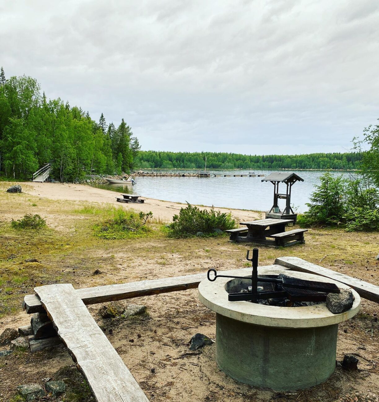 En eldplats med sittbänkar där strand, vatten och skog syns i bakgrunden. Längre bort ser man en brygga som går ut i vattnet, samt en liten motorbåt.