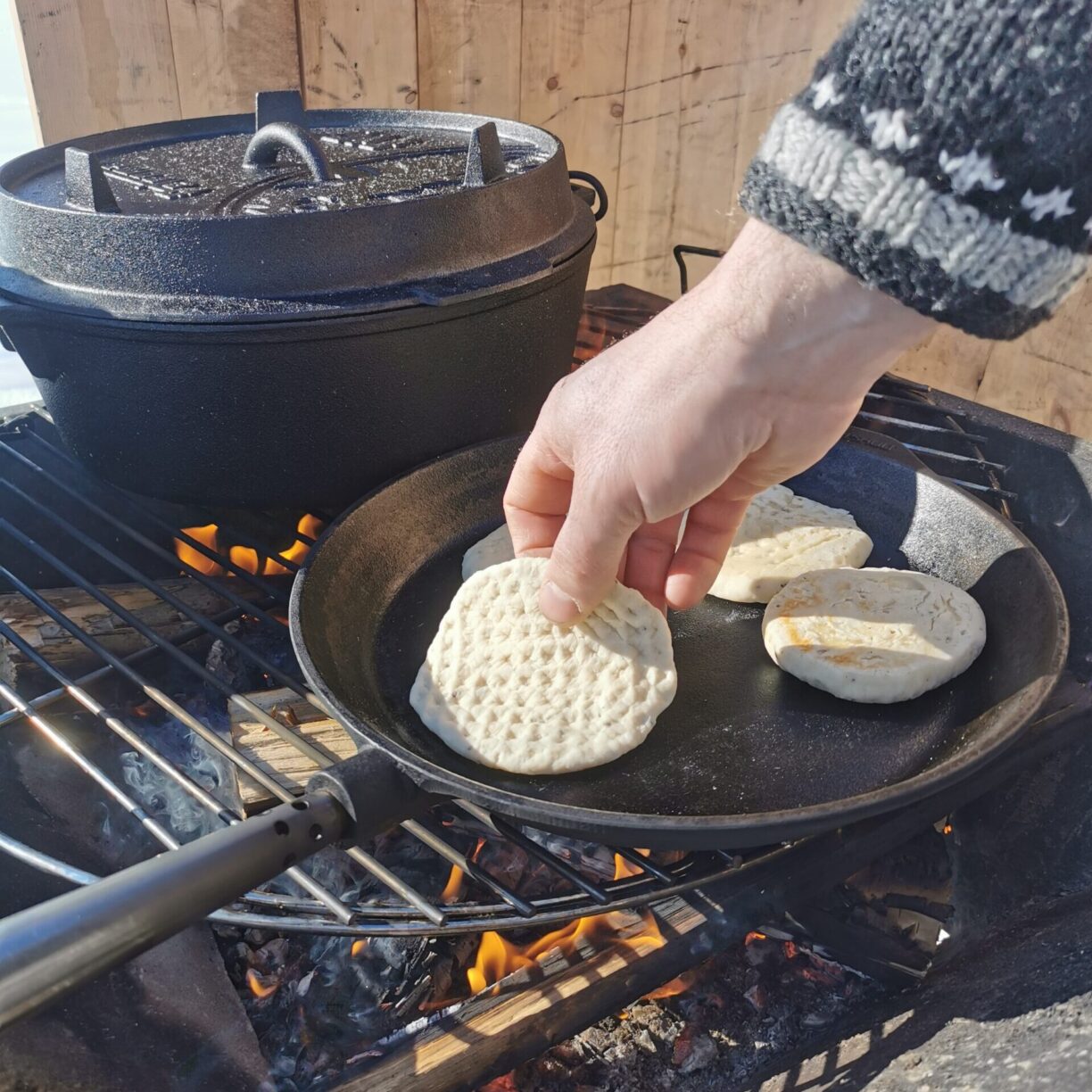 En eld med galler där man ser en stekpanna där en hand vänder en form av bröd.