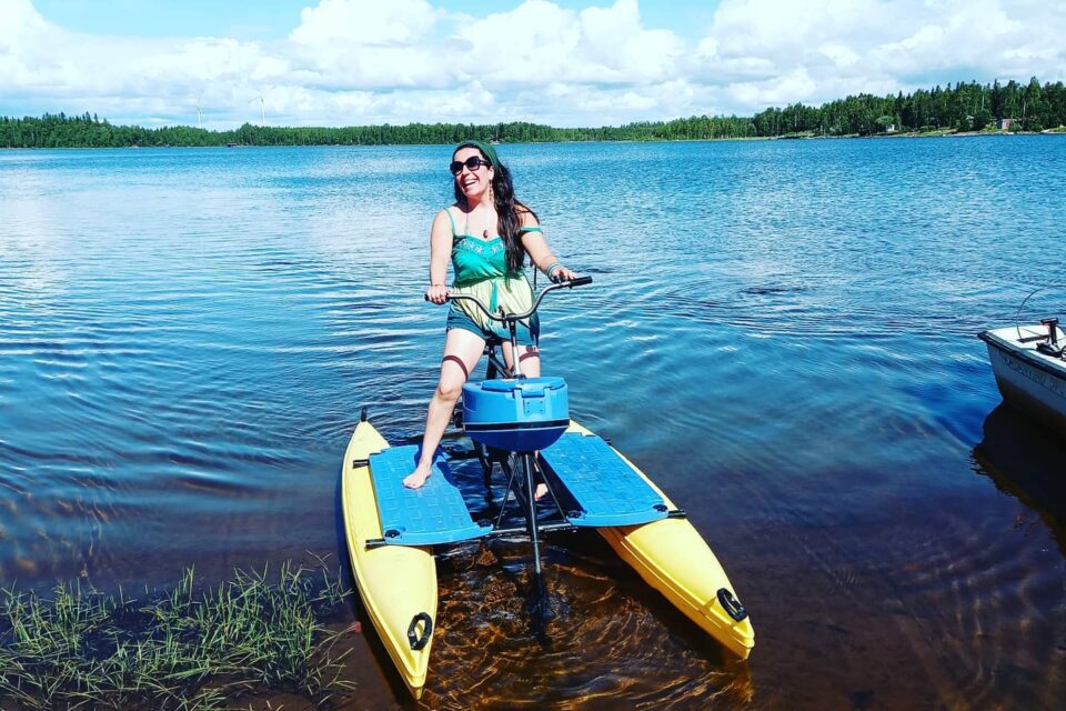 En person står på en hydrobike i vattnet, en cykel på vatten. Blå himmel och i horisonten skymtas en grön skog.