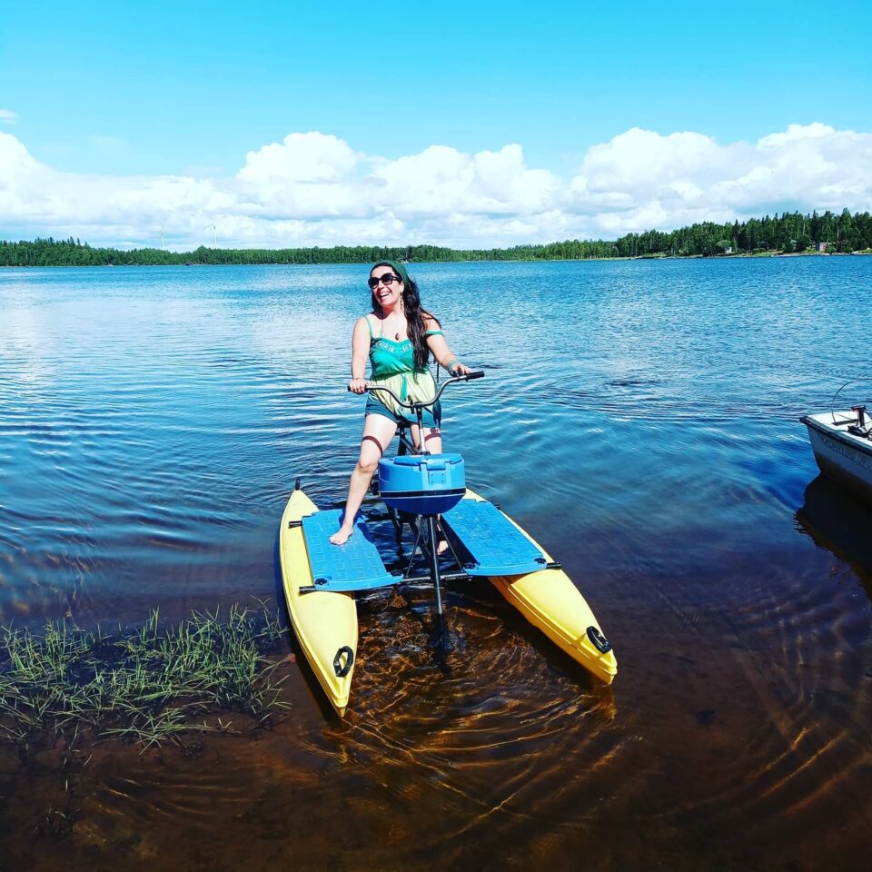 En person står på en hydrobike i vattnet, en cykel på vatten. Blå himmel och i horisonten skymtas en grön skog.