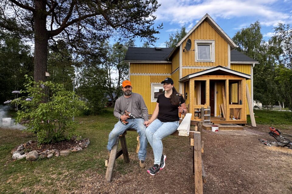 Mimi och Derrick sitter framför sitt gula hus. Till vänster syns lite grönska och ett träd.