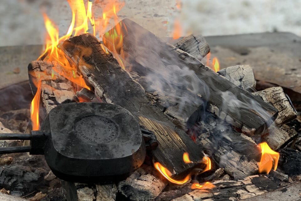 En brasa med gul-orange eldslågor i en grill-ring. Ett smörgåsjärn ligger på elden.