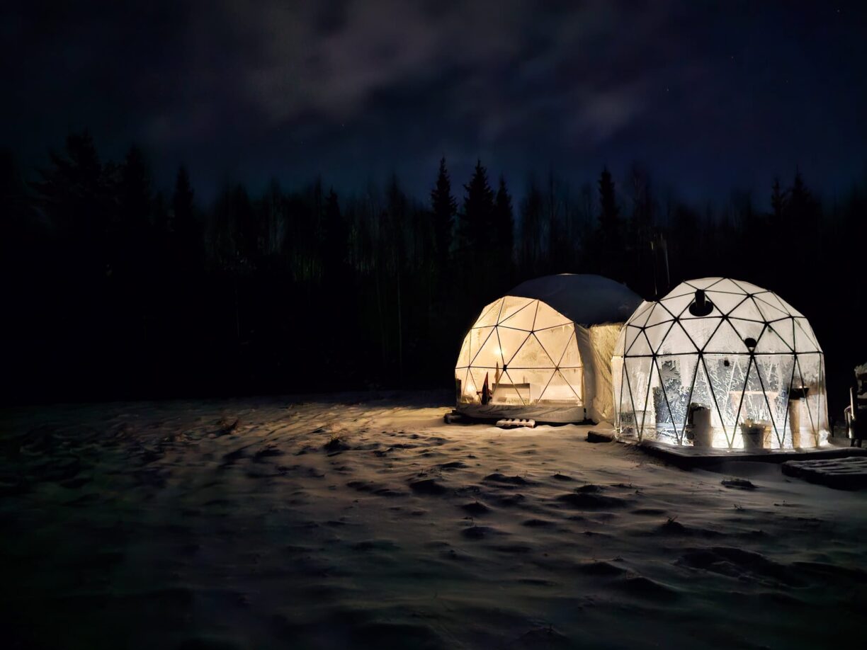 Vit upplyst igloo som står på snötäckt mark. Mörkt runt om hela igloon.