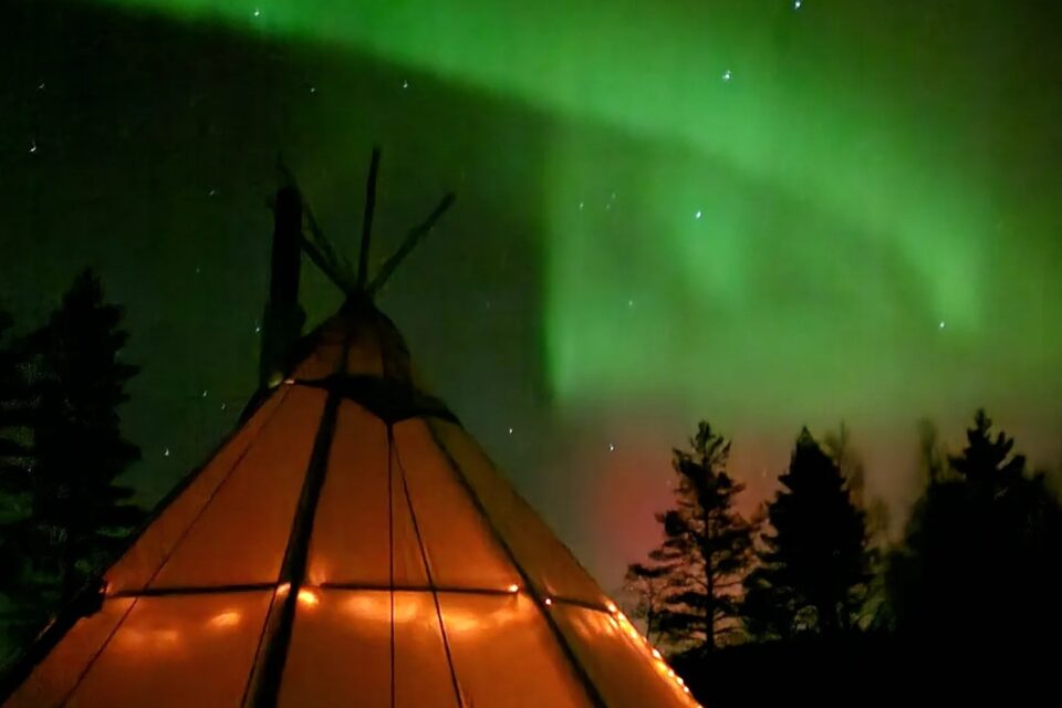 Upplyst kåta mot mörk himmel och norrsken. Mörka granar står i bakgrunden.