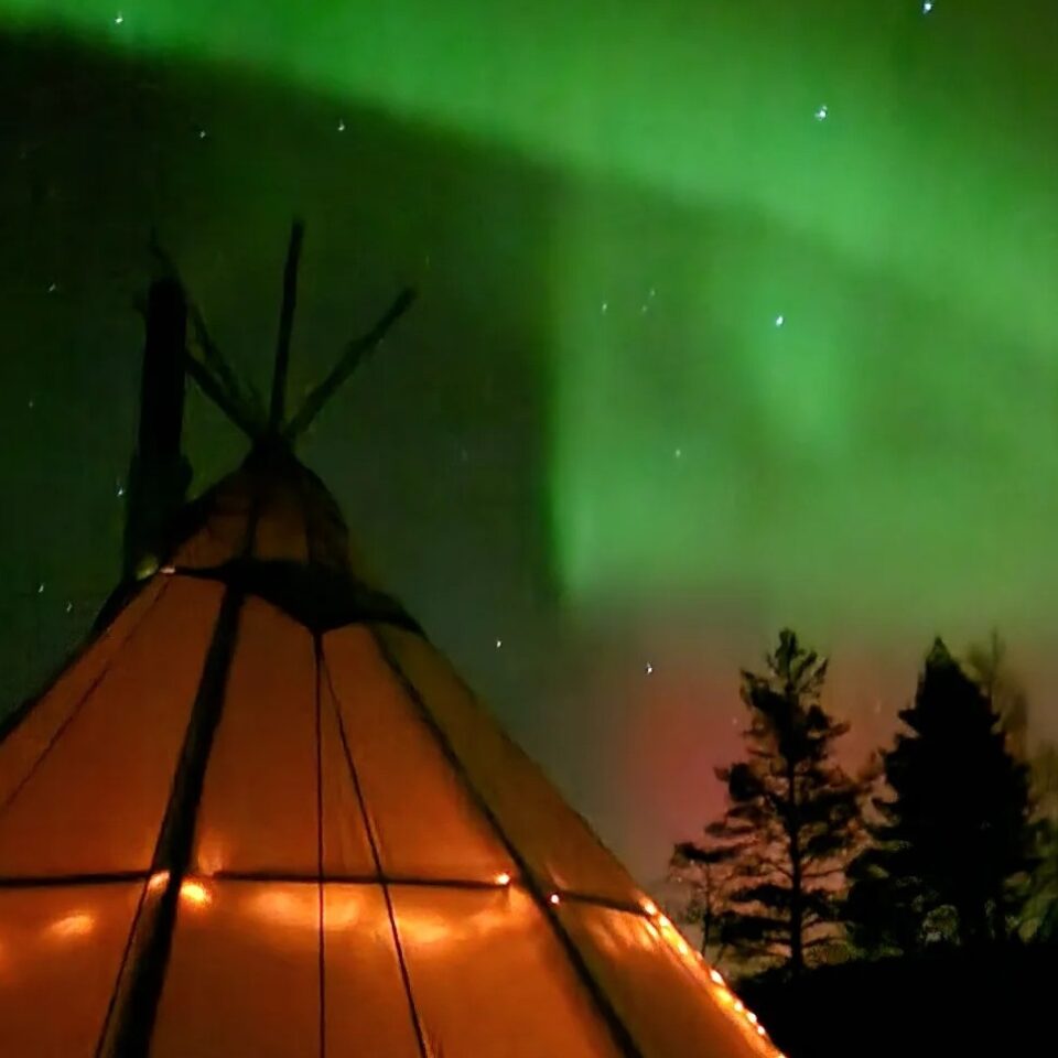 Upplyst kåta mot mörk himmel och norrsken. Mörka granar står i bakgrunden.