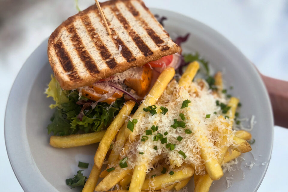 En tallrik med en sandwich fylld med sallad och kyckling, bredvid ligger pommes som är toppad med riven ost och hackad persilja.