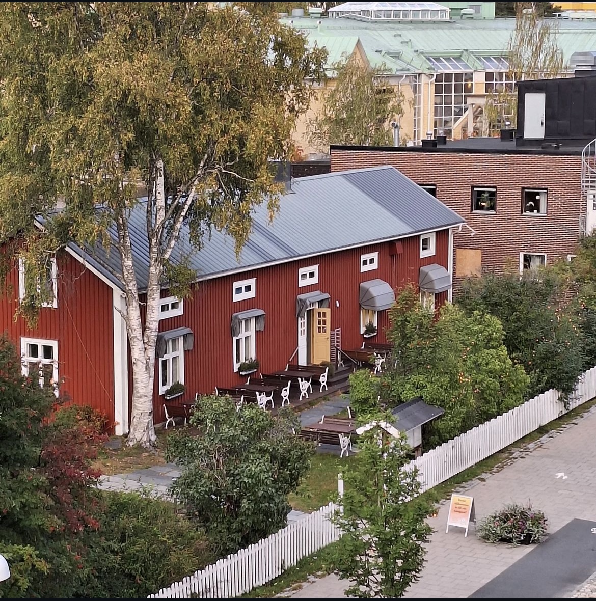 En vybild tagen uppifrån där man ser ett avlångt rött hus med flera fönster och en gul dörr. Framför huset syns grönskande träd och buskar samt ett vitt staket. Framför staketet skymtar en trottoar.