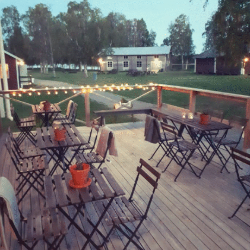 En träaltan med cafébord och stolar i förgrunden, i bakgrunden syns grönt gräs och en grånad lada och gröna träd.
