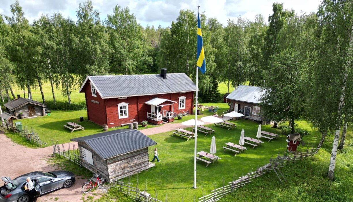 Tre gammal timringar, en röd och två gråa som står på en grönskande tomt med staket runt om. En flaggstång står i mitten och några bänkar och bord står utställda mellan husen på gräset.