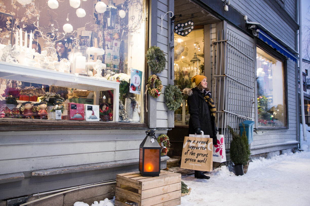 Kvinna i kappa och mössa, med shoppingkassar i händerna, står på en butiksgata i ett vintrigt Kalix.
