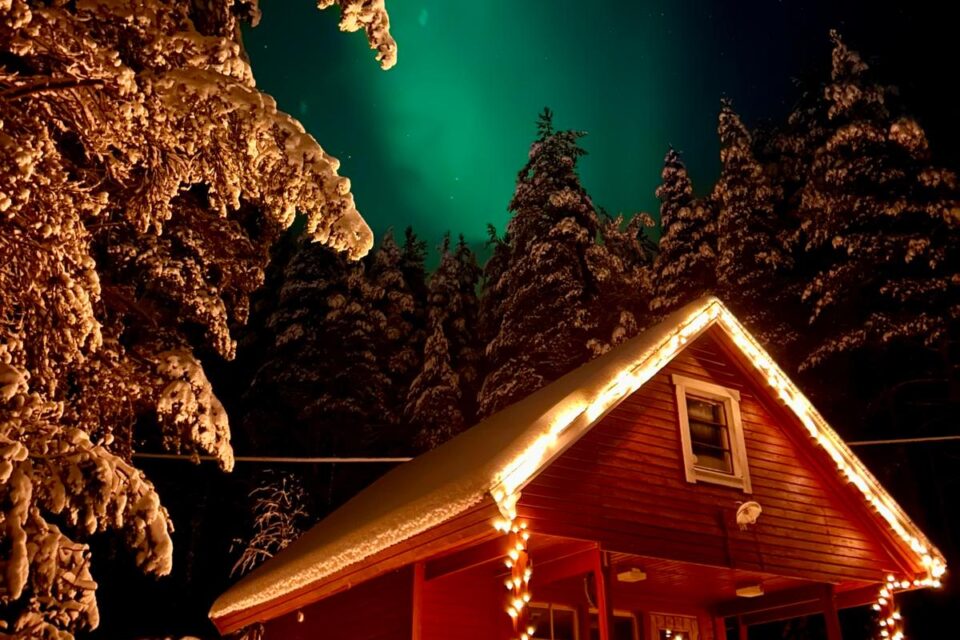 Röd stuga som står inbäddad bland granskog. Stugans farstukvist och nock har pyntats med belysning. Himlen färgas grön från norrskenet ovanför stugan och grantopparna.