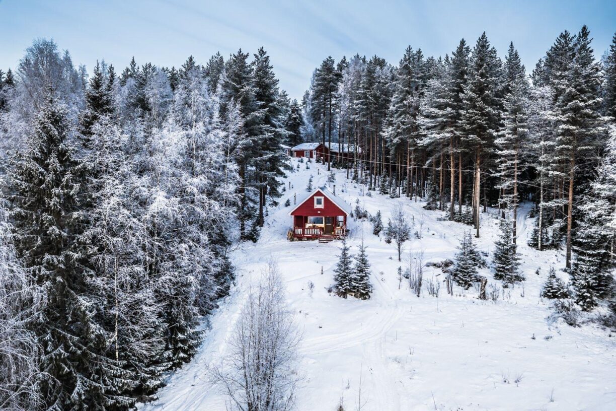 En röd stuga som står på ett snötäckt berg med granskog på båda sidor om stugan.
